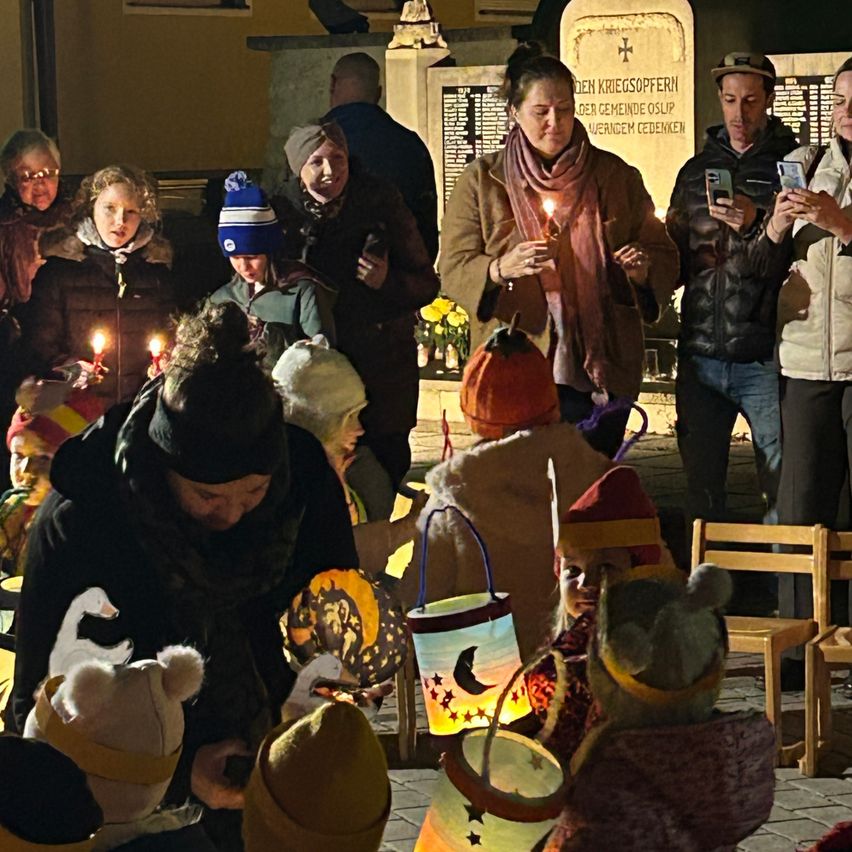 Eine Gruppe von Menschen, einschließlich Erwachsener und Kindern, versammelt sich draußen bei Nacht. Sie halten brennende Kerzen und tragen Winterkleidung. Einige haben Mützen und Schals, während andere Körbe halten. Ein Denkmal ist im Hintergrund.