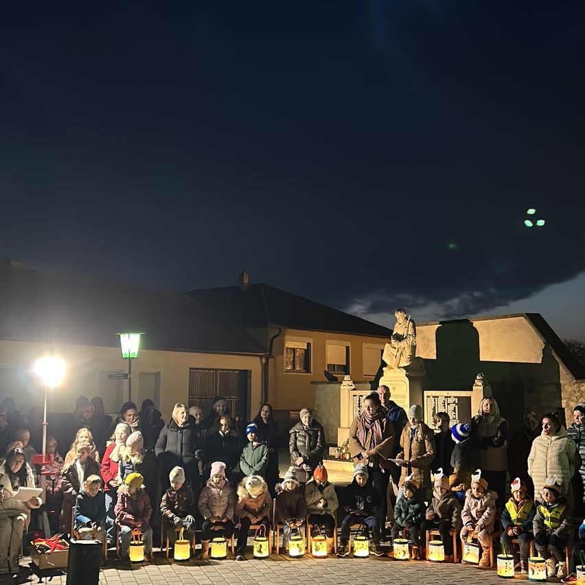 Eine Gruppe von Menschen versammelt sich bei Nacht vor einem Gebäude, einige halten Laternen, während andere auf Stühlen sitzen. Der Himmel ist dunkel und hinter ihnen steht eine Statue.