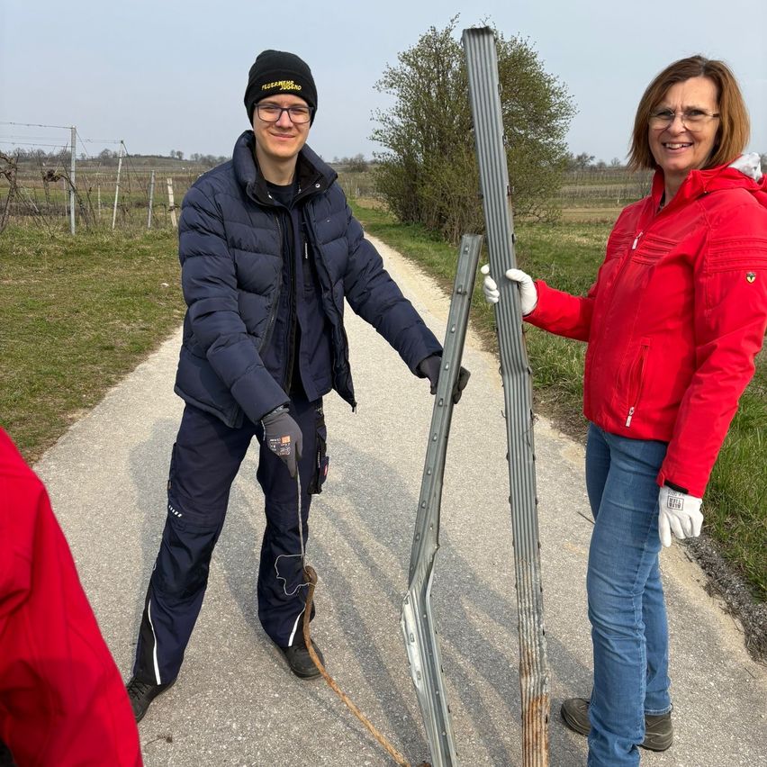 Zwei Personen stehen auf einem Feldweg in einer ländlichen Gegend, wobei eine eine Leiter hält und die andere lächelt. Sie scheinen gemeinsam an einem Projekt zu arbeiten.