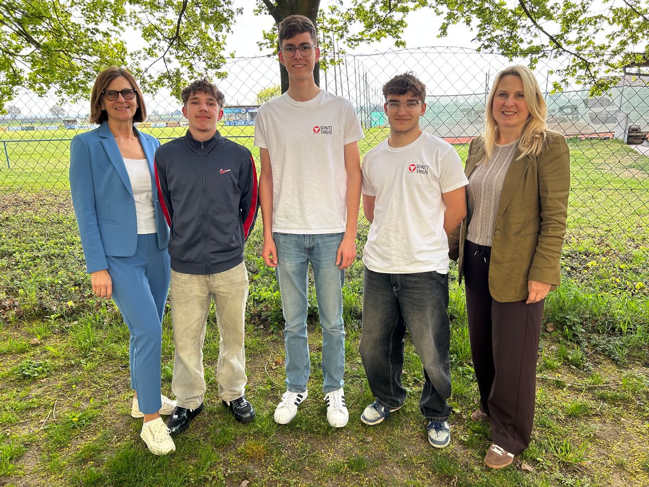 Fünf Personen, darunter drei Jungen und zwei Frauen, stehen auf einem Grasfeld. Hinter ihnen ist ein Metallzaun und ein Sportfeld. Die Jungen tragen Freizeitkleidung.