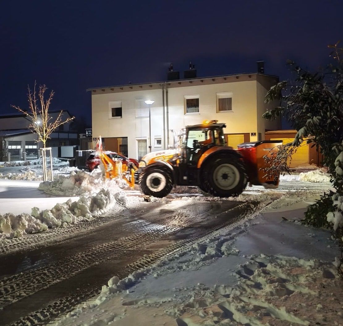 Ein Traktor mit Schneepflug-Aufsatz räumt in der Nacht Schnee von einer Straße vor einem Gebäude.