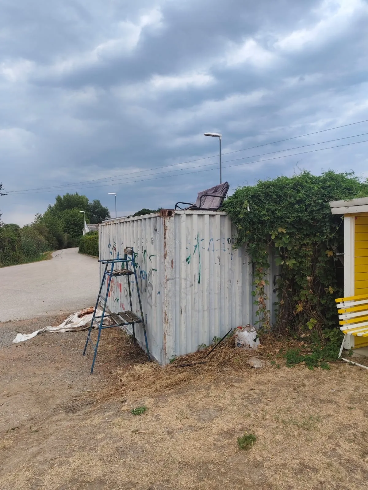 Ein Metalllagercontainer mit Graffiti darauf steht auf einem Feldweg. Ein Stuhl ist darauf.
