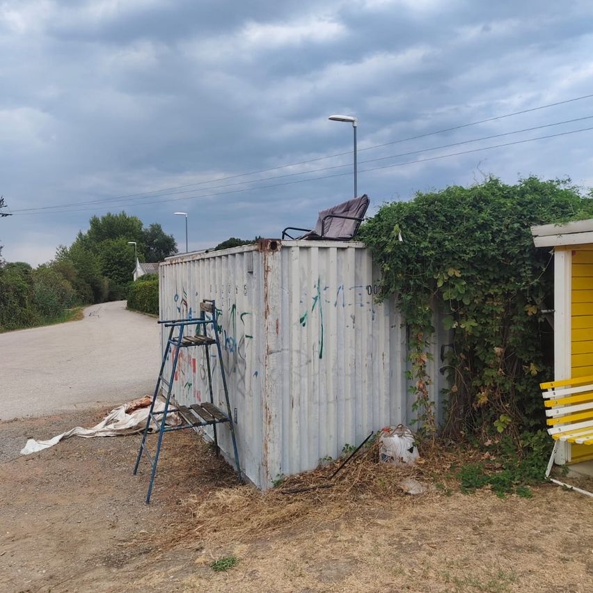Ein Metalllagercontainer mit Graffiti darauf steht auf einem Feldweg. Ein Stuhl ist darauf.