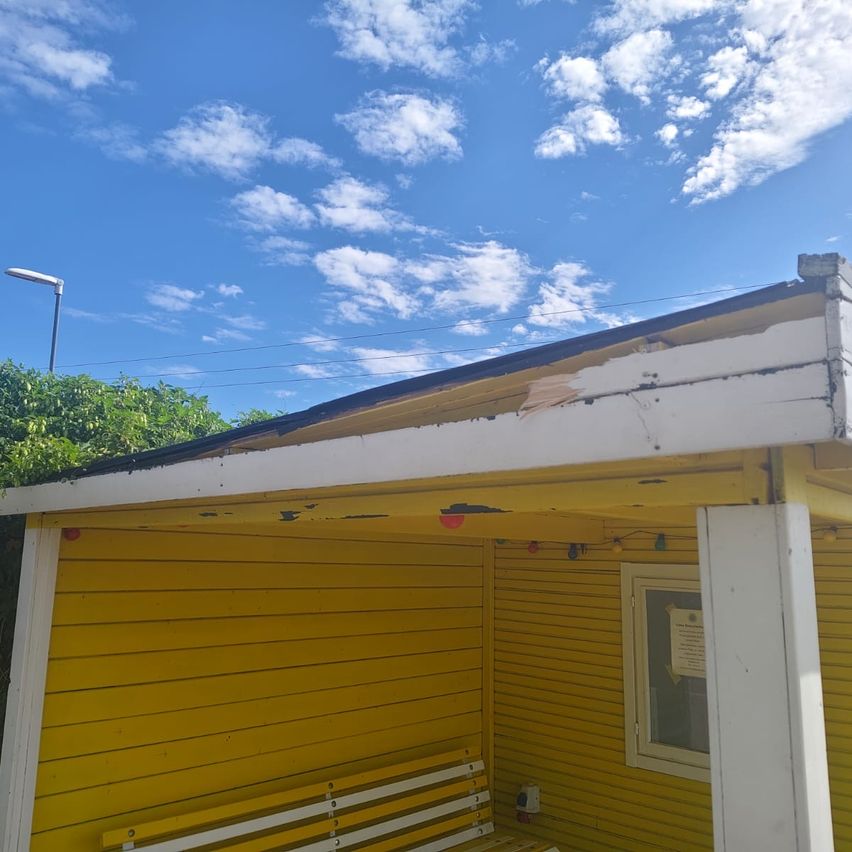 Ein gelbes Holzgebäude mit beschädigtem Dach und einer Bank davor. Der Himmel ist blau mit verstreuten Wolken.