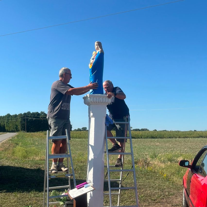 Zwei Männer stehen auf Leitern, einer hält eine blaue Statue einer Frau, während der andere sie putzt. Sie befinden sich in einem Grasfeld mit klarem blauem Himmel.