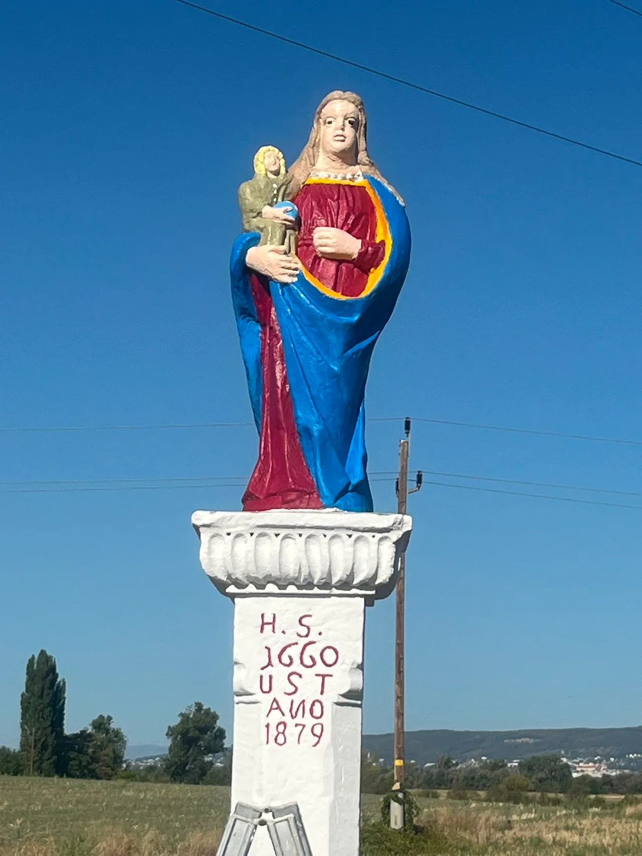 Eine große Statue einer Frau, die ein Kind hält, in Blau und Rot bemalt, steht auf einem Podest mit dem Text 'H.S. 1660 U.S.T. 1879'. Die Statue steht vor einem klaren blauen Himmel mit einigen Bäumen und Stromleitungen im Hintergrund.