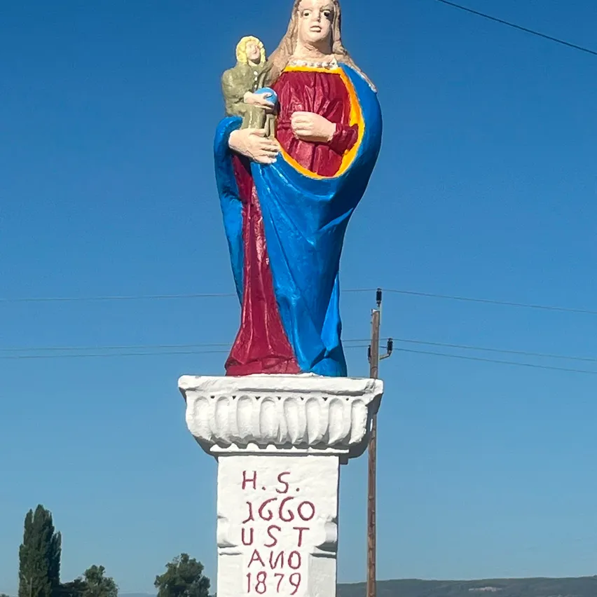 Eine große Statue einer Frau, die ein Kind hält, in Blau und Rot bemalt, steht auf einem Podest mit dem Text 'H.S. 1660 U.S.T. 1879'. Die Statue steht vor einem klaren blauen Himmel mit einigen Bäumen und Stromleitungen im Hintergrund.