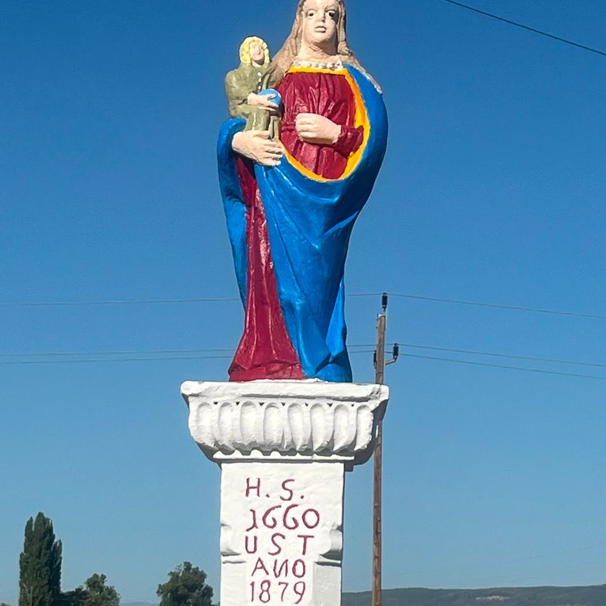 Eine große Statue einer Frau, die ein Kind hält, in Blau und Rot bemalt, steht auf einem Podest mit dem Text 'H.S. 1660 U.S.T. 1879'. Die Statue steht vor einem klaren blauen Himmel mit einigen Bäumen und Stromleitungen im Hintergrund.
