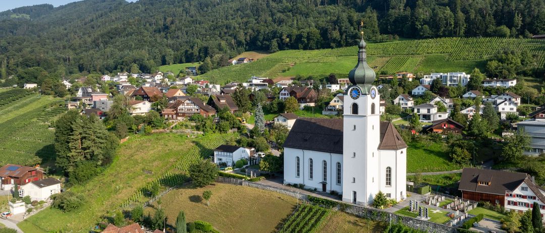 Ein Luftbild einer Kirche mit einem grünen Turm und Uhr, eingebettet in einem Tal, umgeben von Weinbergen und Häusern. Im Hintergrund sind Berge zu sehen.