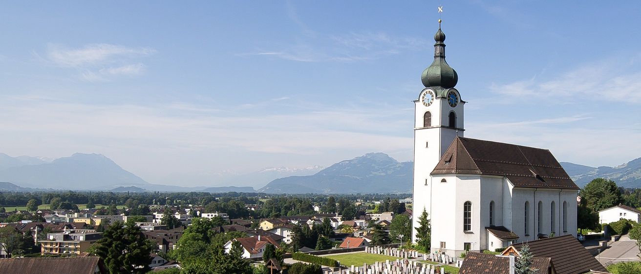 Auf einem Hügel steht eine Kirche mit einer grünen Kuppel und einem Glockenturm. Die Kirche ist von einem Friedhof umgeben, mit Bergen in der Ferne.