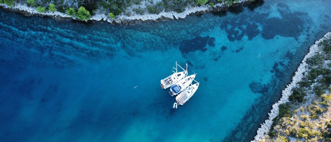 Luftaufnahme von drei Booten, die in einer ruhigen Bucht mit klarem blauem Wasser und Felsküste verankert sind.