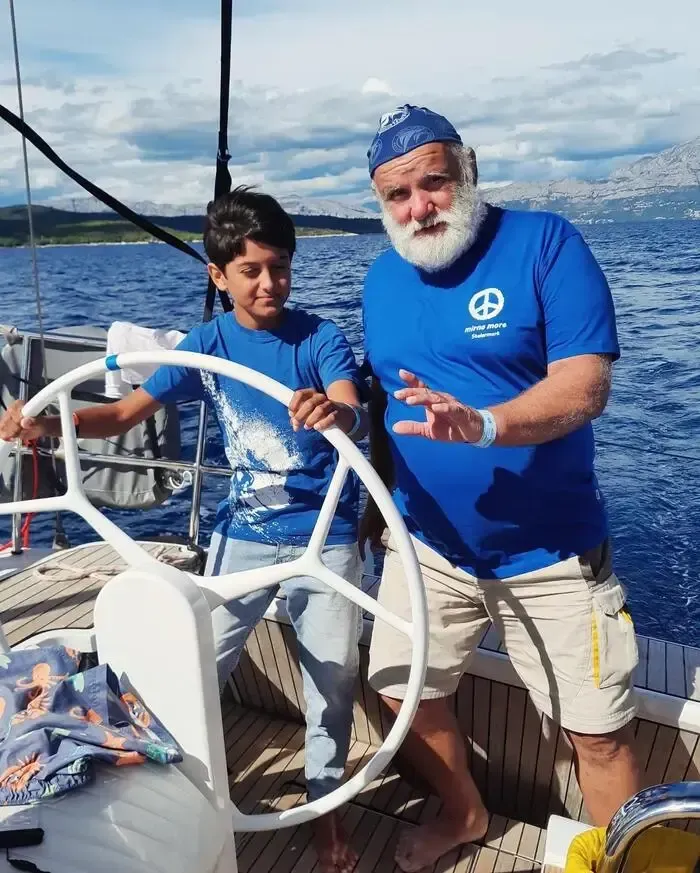Ein älterer Mann und ein Junge sind auf einem Boot, der Junge steuert und der Mann erklärt etwas. Sie tragen beide blaue T-Shirts.