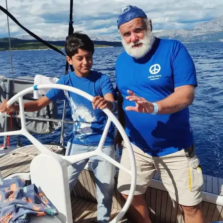 Ein älterer Mann und ein Junge sind auf einem Boot, der Junge steuert und der Mann erklärt etwas. Sie tragen beide blaue T-Shirts.