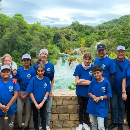 Eine Gruppe von Menschen in blauen T-Shirts mit einem Friedenssymbol posiert vor einem Wasserfall und einer Steinmauer.