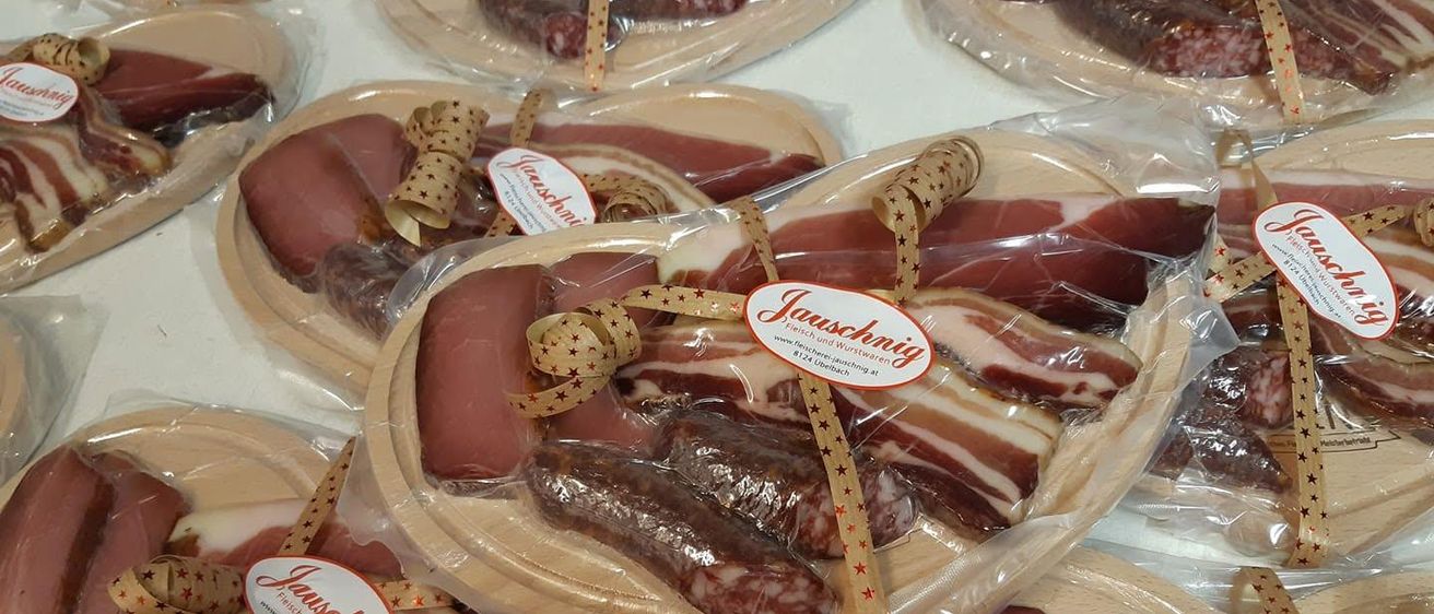 Eine Sammlung von Holztabletts, die verschiedene Arten von Wurstwaren enthalten, in Plastik verpackt und mit dekorativen Bändern verziert. Die Wurstsorten umfassen Scheiben von Speck, Salami und anderen Wurstsorten.