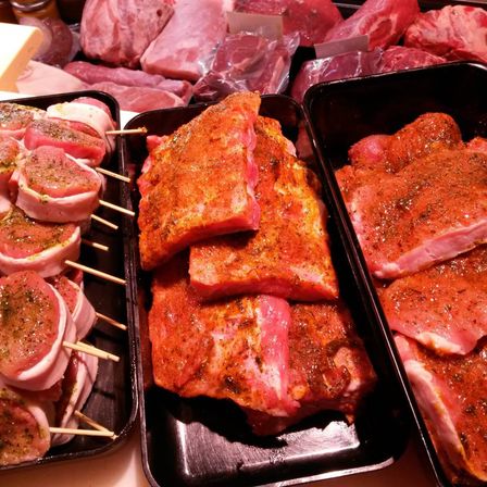 Eine Nahaufnahme verschiedener Fleischstücke, einschließlich marinierter Rippen, gefüllter Fleischspieße und geschnittenem Schweinefleisch, die auf Tabletts in einem Fleischgeschäft ausgestellt sind.