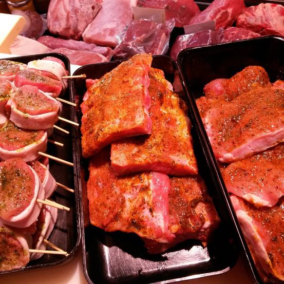 Eine Nahaufnahme verschiedener Fleischstücke, einschließlich marinierter Rippen, gefüllter Fleischspieße und geschnittenem Schweinefleisch, die auf Tabletts in einem Fleischgeschäft ausgestellt sind.