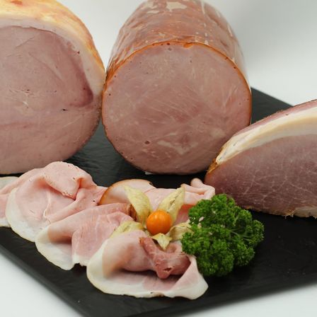 Nahaufnahme verschiedener rosa Schinkenstücke auf einem schwarzen Schieferteller, einschließlich Scheiben und einem ganzen Stück, garniert mit Petersilie und einer kleinen Orange.