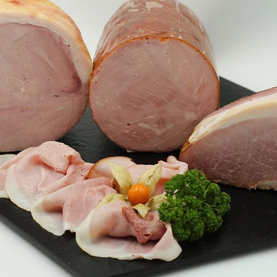Nahaufnahme verschiedener rosa Schinkenstücke auf einem schwarzen Schieferteller, einschließlich Scheiben und einem ganzen Stück, garniert mit Petersilie und einer kleinen Orange.