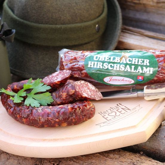 Ein Holzschneidebrett mit Scheiben von Ubelbacher Hirschsalami-Wurst, garniert mit Petersilie, und einem Messer. Ein grüner Hut und eine Flasche sind im Hintergrund.