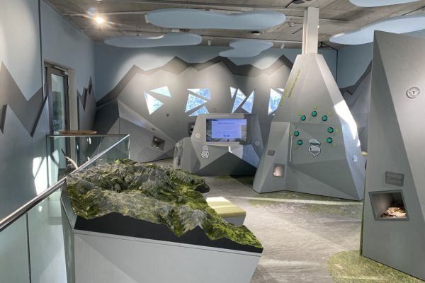 Ein Innenbereich mit grauen geometrischen Formen und grünem moosbewachsenem Gelände. Ein Monitor steht auf dem Schreibtisch, und links befindet sich eine Glasbrüstung.
