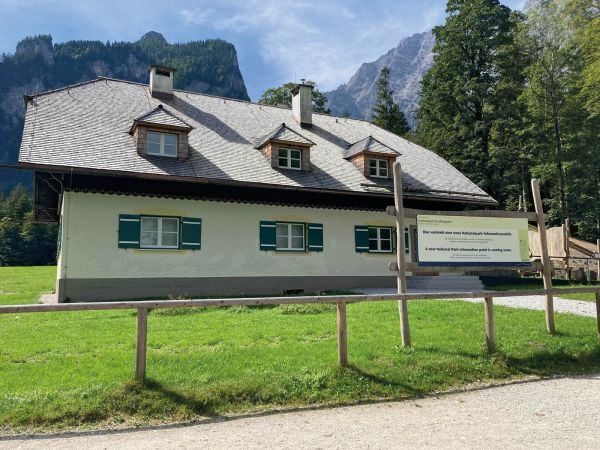Ein kleines Haus mit grauem Dach und grünen Fensterläden steht auf einer Rasenfläche mit einem Holzzaun. Vor dem Haus befindet sich ein Schild.