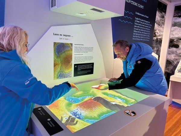 Zwei Personen, die blaue Jacken tragen, betrachten eine interaktive Karte auf einem Tisch in einem Museum. Eine Frau auf der linken Seite zeigt auf die Karte, während der Mann auf der rechten Seite sie ansieht. Hinter ihnen befindet sich eine Wand mit einem Informationsschild.