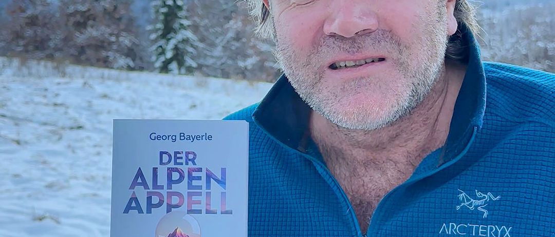 Ein Mann steht in einer verschneiten Landschaft und hält ein Buch mit dem Titel 'Der Alpen Appell'. Das Cover zeigt eine Sanduhr mit einem Berggipfel. Er trägt eine blaue Arcteryx-Jacke.