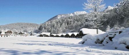 Eine verschneite Landschaft mit einem Bergrücken im Hintergrund und einigen schneebedeckten Hütten im Vordergrund.