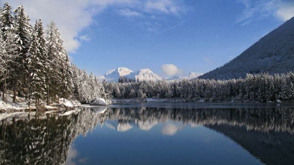 Eine ruhige Winterlandschaft mit schneebedeckten Kiefern, einem ruhigen See, der den Himmel und die Berge im Hintergrund reflektiert.