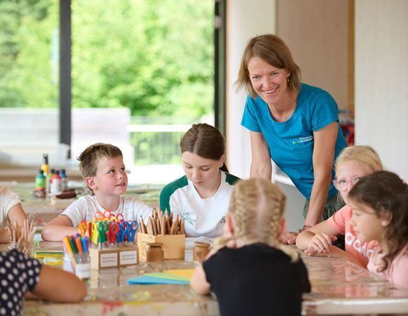 Eine Gruppe von Kindern sitzt mit verschiedenen Bastelutensilien am Tisch, eine lächelnde Frau steht hinter ihnen.