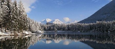 A serene winter landscape with a calm lake reflecting snow-covered pine trees and distant mountains under a blue sky with some clouds.