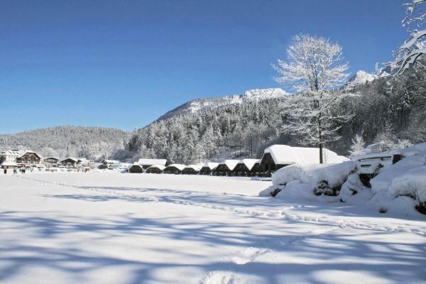 Eine Winterszene zeigt eine verschneite Landschaft mit einer Reihe von Chalets und schneebedeckten Bäumen. Berge sind in der Ferne zu sehen.