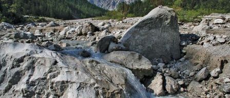 A stream of water flows through rocky terrain, surrounded by lush green trees and a backdrop of mountains under a clear sky.
