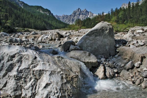 Ein Wasserstrom fließt durch felsiges Gelände, umgeben von üppigem Grün der Bäume und mit einem Hintergrund aus Bergen unter einem klaren Himmel.
