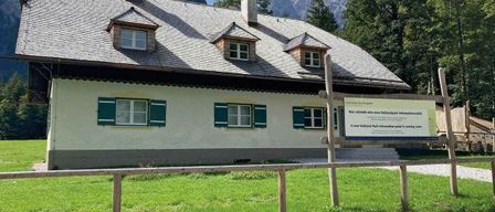 A small white house with green shutters and two chimneys in a grassy field with mountains in the background.