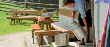 A man pours milk from a silver jug into a large metal pot on the porch of a house with wooden benches and a fence.