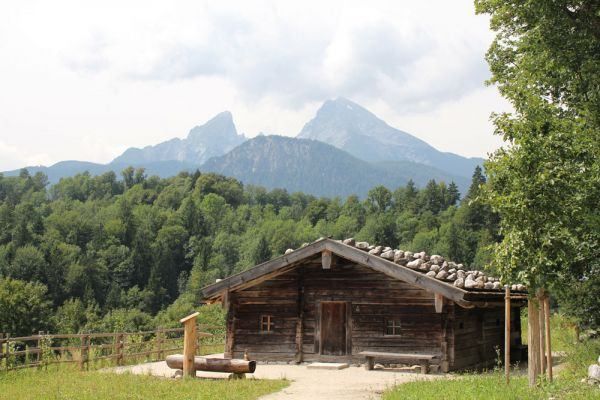 Eine rustikale Holzhütte mit einem Steindach befindet sich in einem üppigen grünen Tal, umgeben von einem Holzzaun und einer Bank, mit majestätischen Bergen im Hintergrund.