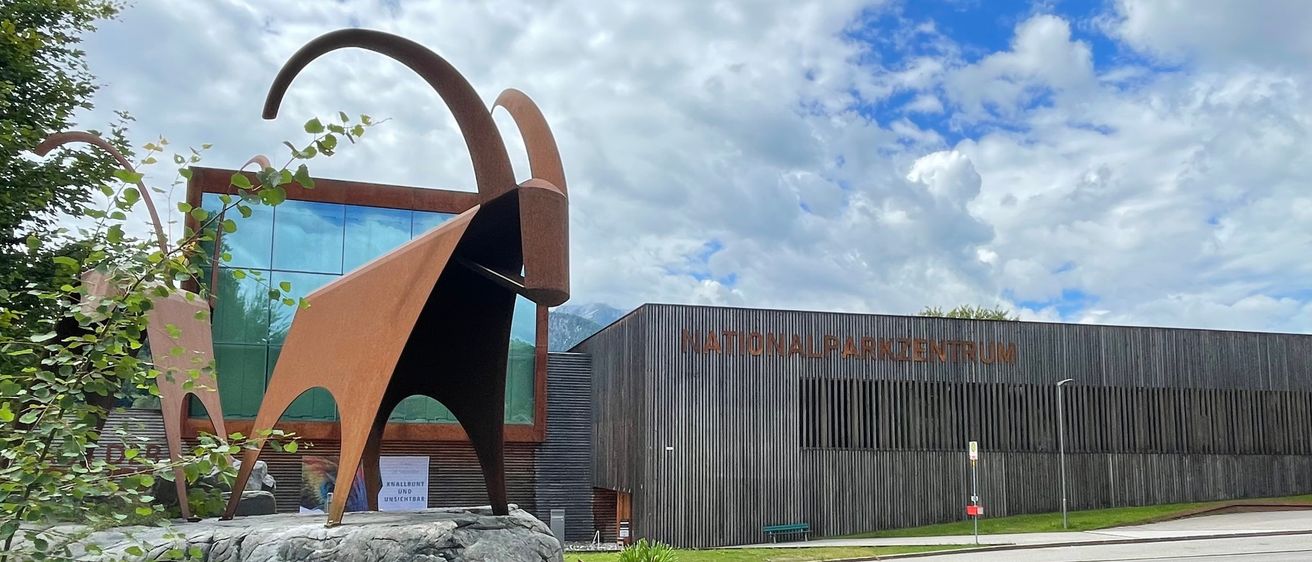 Eine Ziegenstatue steht vor einem modernen Gebäude mit Glaswänden und einem Schild, auf dem 'NATIONALPARKZENTRUM' steht. Die Szenerie ist vor einem Hintergrund von Bergen und bewölktem Himmel angesiedelt.