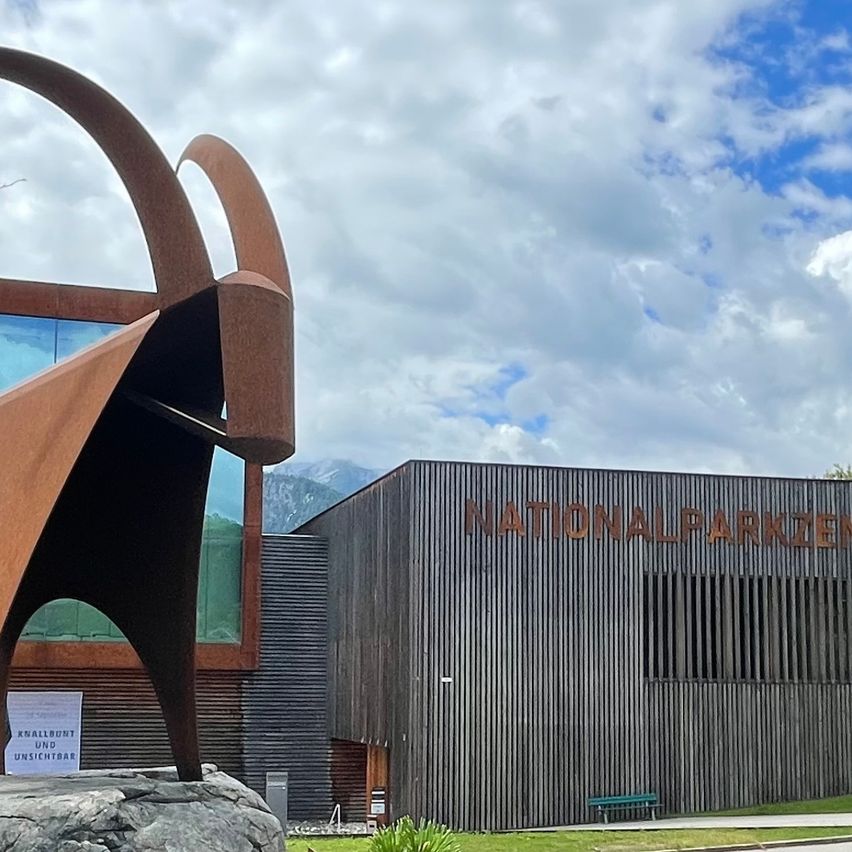 Eine Ziegenstatue steht vor einem modernen Gebäude mit Glaswänden und einem Schild, auf dem 'NATIONALPARKZENTRUM' steht. Die Szenerie ist vor einem Hintergrund von Bergen und bewölktem Himmel angesiedelt.