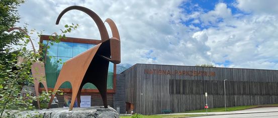 A goat sculpture stands before a modern building with glass walls and a sign reading 'NATIONALPARKZENTRUM'. The scene is set against a backdrop of mountains and cloudy skies.