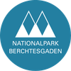 Das Logo für Nationalpark Berchtesgaden. Ein blauer Kreis mit drei weißen Dreiecken vertikal gestapelt und der Name in weiß geschrieben.