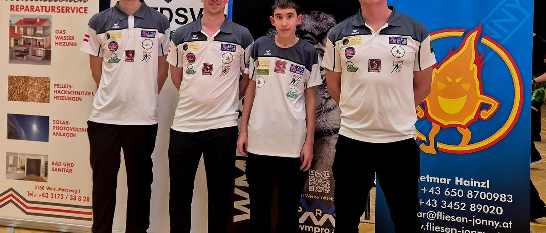 Vier Personen stehen in einer Reihe in einem Raum. Sie tragen alle weiße und graue Shirts. Sie posieren für ein Foto. Hinter ihnen befinden sich zwei Banner, eines mit dem Wort FLIESEN-JOHN und das andere mit einem QR-Code.