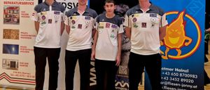 Vier Personen stehen in einer Reihe in einem Raum. Sie tragen alle weiße und graue Shirts. Sie posieren für ein Foto. Hinter ihnen befinden sich zwei Banner, eines mit dem Wort FLIESEN-JOHN und das andere mit einem QR-Code.