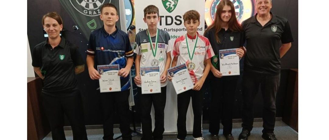 A group of young dart players, four boys and two girls, are standing in front of a dartboard banner and holding certificates and medals. They are wearing matching black and white uniforms.