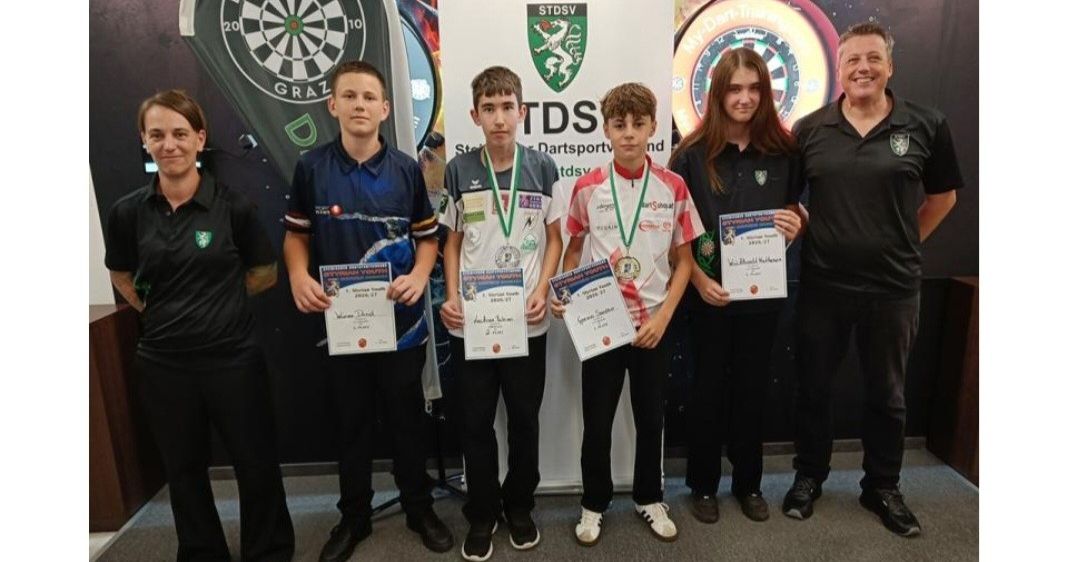 Eine Gruppe junger Dartspieler, vier Jungen und zwei Mädchen, stehen vor einem Dartboard-Banner und halten Urkunden und Medaillen in den Händen. Sie tragen passende schwarze und weiße Uniformen.