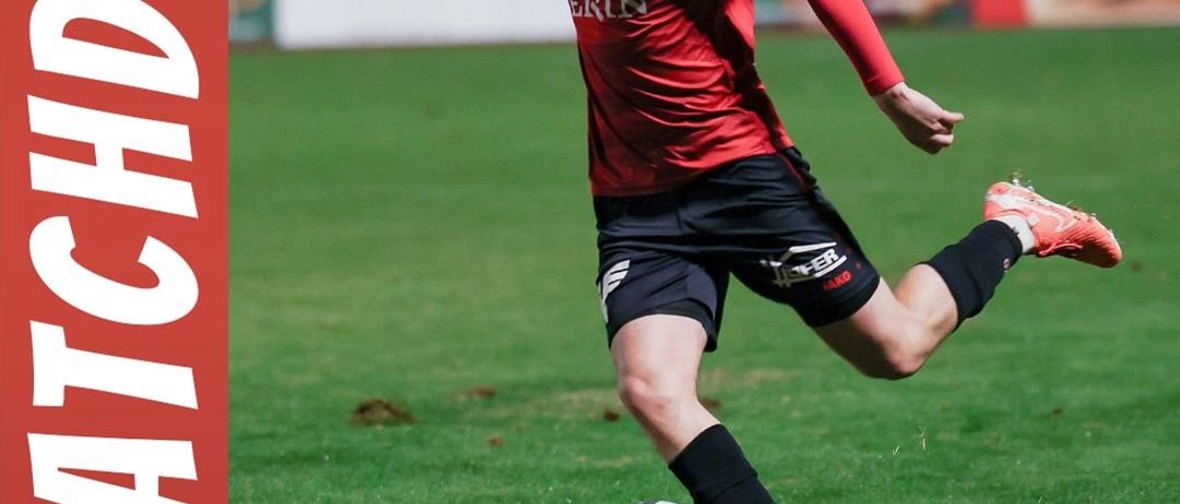 Ein Fußballspieler in einem roten Trikot tritt den Ball während eines Spiels. Der Spieler trägt rote Turnschuhe und Shorts. Der Platz hat weiße Markierungen. Unten befinden sich zwei Logos, eines mit dem Text SC Gerin Druck Wolkersdorf und das andere mit VS.