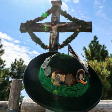Ein traditioneller bayerischer Hut mit verschiedenen Verzierungen steht vor einem Kruzifix unter einem blauen Himmel. Das Kruzifix ist mit einer Girlande geschmückt, und die Szenerie wird von immergrünen Bäumen eingerahmt.