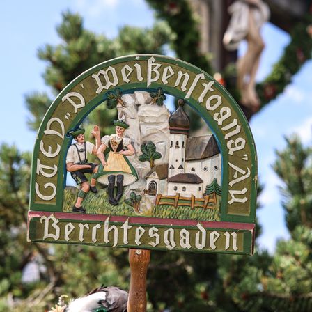 Ein Schild, das zwei Personen in traditioneller bayerischer Kleidung vor einem Haus mit Turm zeigt. Das Schild trägt die Aufschrift 'Gedenk' und 'Berchtesgaden'.