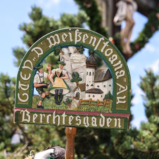 Ein Schild, das zwei Personen in traditioneller bayerischer Kleidung vor einem Haus mit Turm zeigt. Das Schild trägt die Aufschrift 'Gedenk' und 'Berchtesgaden'.